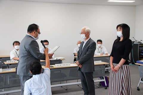 読み上げるスーツの男性と、対面する男性と女性の二人の写真
