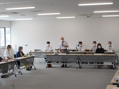 立ち上がり、マイクで話しているワイシャツの男性と他の参加者たちの写真