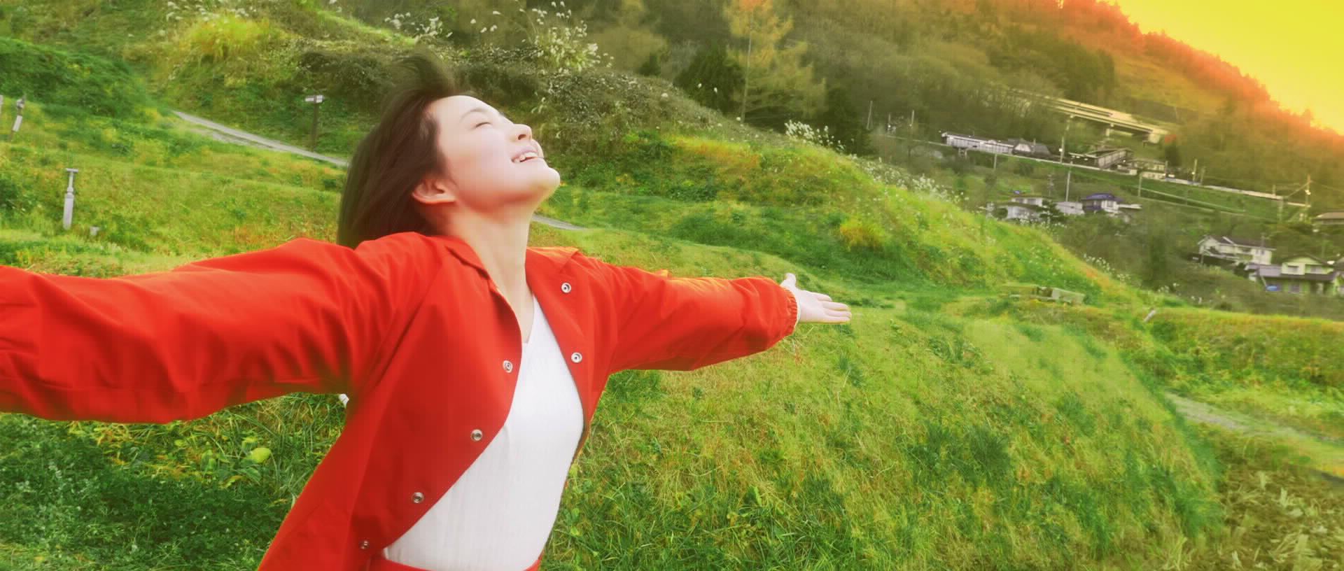 里山の風景を背に、赤と白の服を着た女性が両手を広げてほほ笑んでいる写真