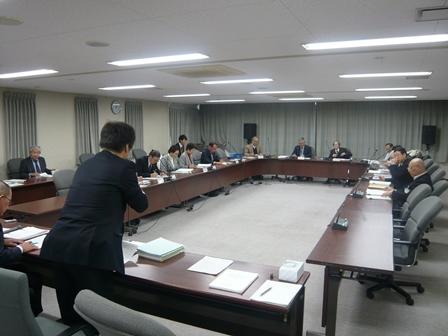 会議室でスーツの男性が前に立って、他の人々は座って協議をしている写真