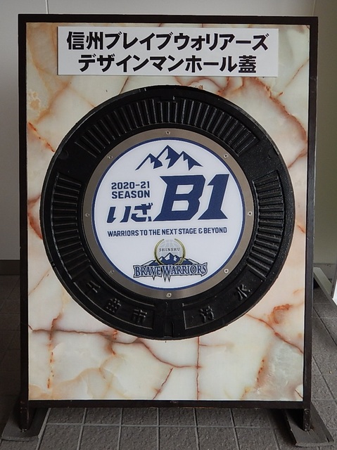 「信州ブレイブウォリアーズデザインマンホール蓋」とと書かれた張り紙の下に、「いざ、B1」と書かれたマンホールが埋め込まれている台座の写真