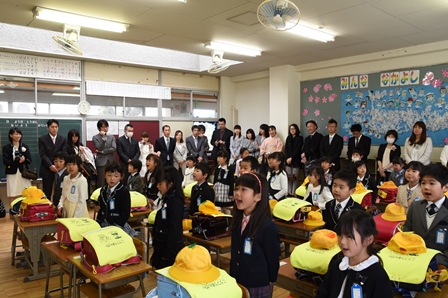教室内で、新しいランドセルと学生帽が置かれた机を前に立つ児童達と、それを見守る保護者達の写真