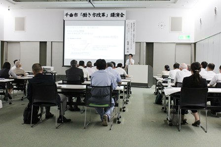 スクリーンに資料を映しながら講師が話し、参加者らは着席して受講している写真
