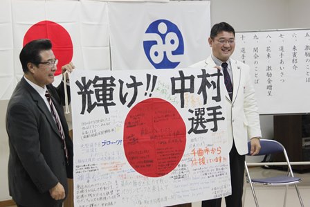 輝け!中村選手という大きな字とともにたくさんの寄せ書きが書かれている日の丸国旗を持っている白いスーツ姿の中村選手と市長