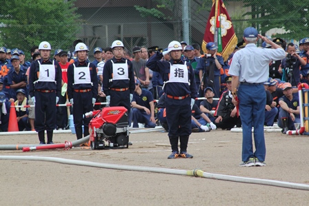 消防ポンプ操法大会で入賞した選手4名と表彰者のグラウンドでの写真