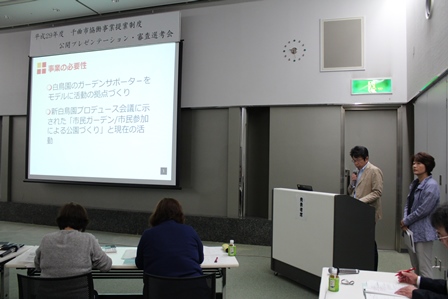 スクリーンを使いマイクでプレゼンテーションをしている男性の写真
