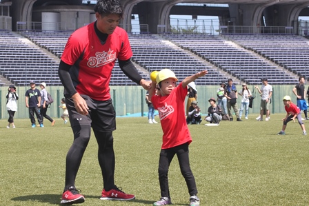 スタジアムの芝鵜の上で赤いユニフォームを着た男の子が指導を受けながらボールを投げている写真