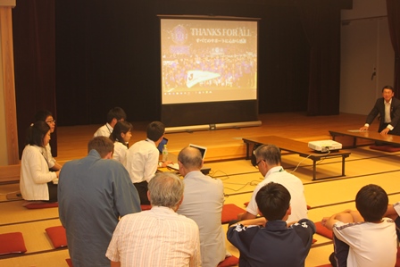 畳の部屋でスクリーンに映った活動報告を見ている大学生5人と市の職員