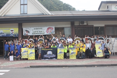 応援バナーを掲げたバスケットボールチームと市民が駅前で集合し元気に映っている集合写真