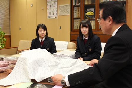 介護用パジャマを机の上に広げている岡田市長と、それを見つめる制服姿の女子生徒2人の写真