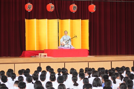 中学校の体育館のステージで三味線漫談をする着物を着た漫談家の女性の写真