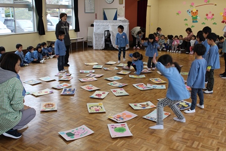 A4サイズはある大きな絵札を立って囲んで保育士さんとカルタをしている子供たちの写真