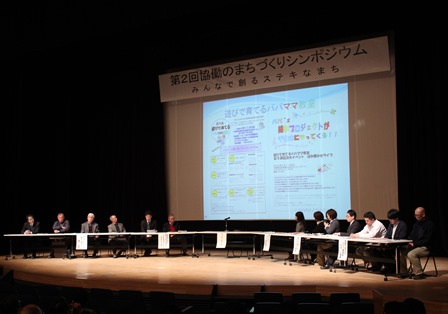 大きなスクリーンのあるステージ上に半円状に並べられた長机に座る参加者たちの写真