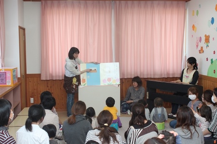 紙芝居のようなものを読む職員とピアノを弾く職員、それを聞く約15組の親子の写真