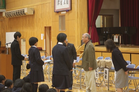 中学校体育館で冊子の贈呈を行う団体のメンバーと受け取る中学生3名の写真