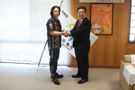 千曲市の市章の旗の前で握手する市長とダーツチームの松林さんの写真