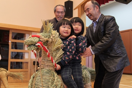 藁で作られた馬に乗るおかっぱの女の子二人とそれを支える老人男性の写真