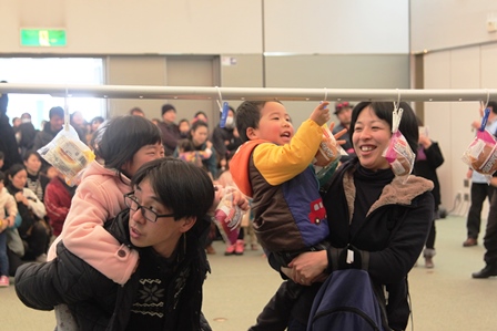 子供を抱っこやおんぶでパイプに吊るされた菓子パンを取らせる親子の写真