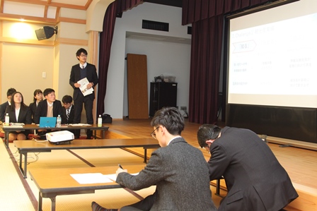 温泉旅館の宴会場のような場所のスクリーンで発表する学生たちの写