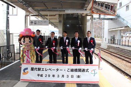 駅のホームで着ぐるみのキャラクターと一緒に開通式を行う関係者一同の写真