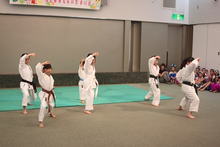 会館内でスペースで空手の型を発表する子供たちと、後ろで出番を待つチアダンスの子供たちの写真