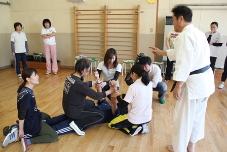 不審者役の男性を複数人で抑え込む保育士達と、指導する道着姿の男性の写真