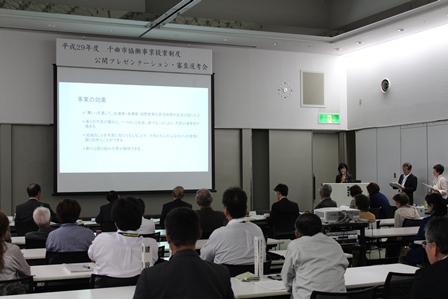会議室でスクリーンを使い大勢の前でプレゼンテーションをしている最中の写真