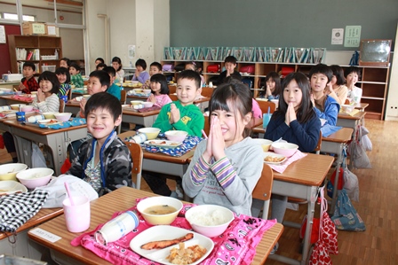 机に置いた給食を前に、両手を合わせいただきますのポーズをとる児童達の写真