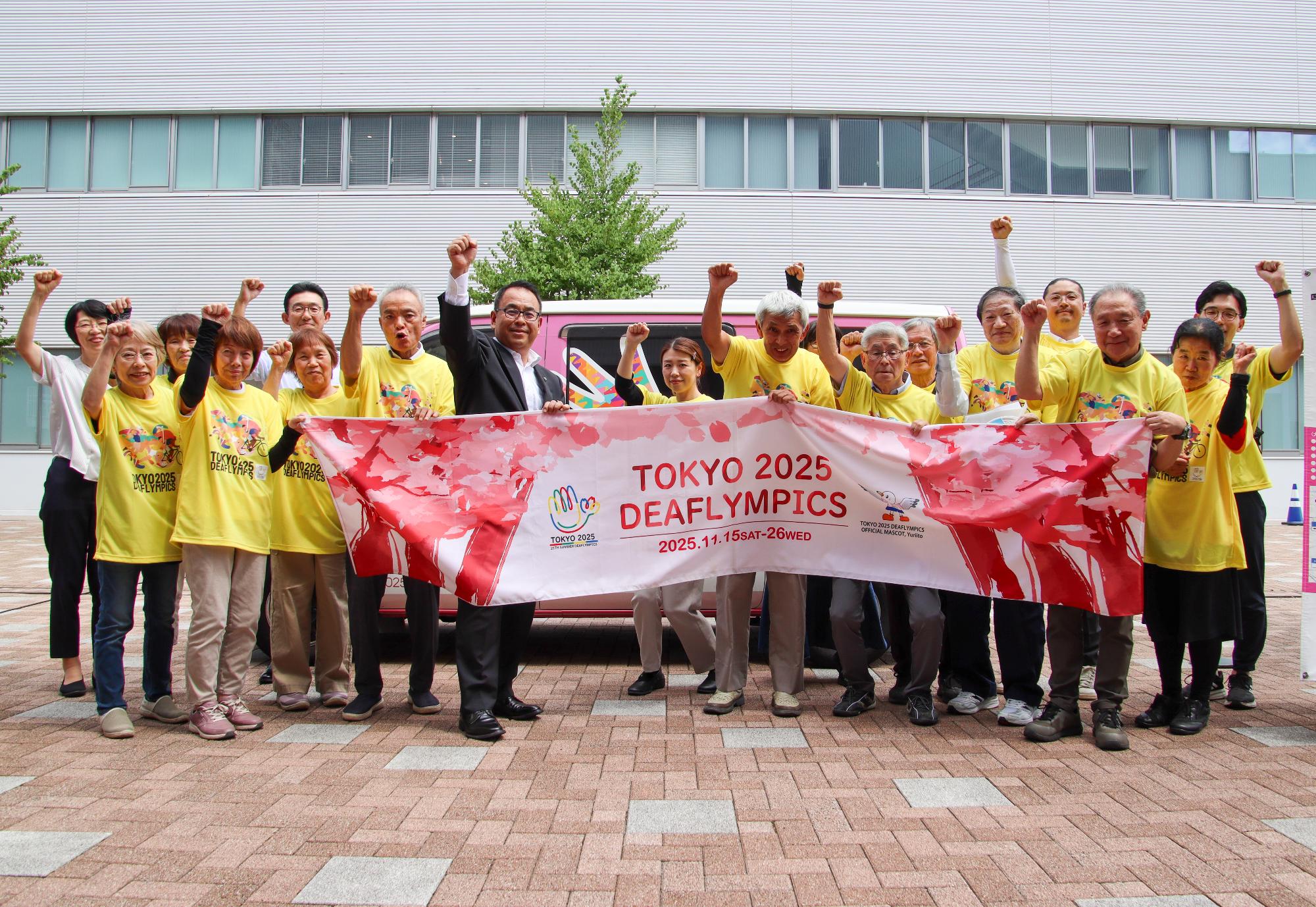 キャラバンカーの前で東京2025デフリンピックの横断幕を持ち、こぶしを上げる小川市長とデフリンピックキャラバンカーのスタッフ
