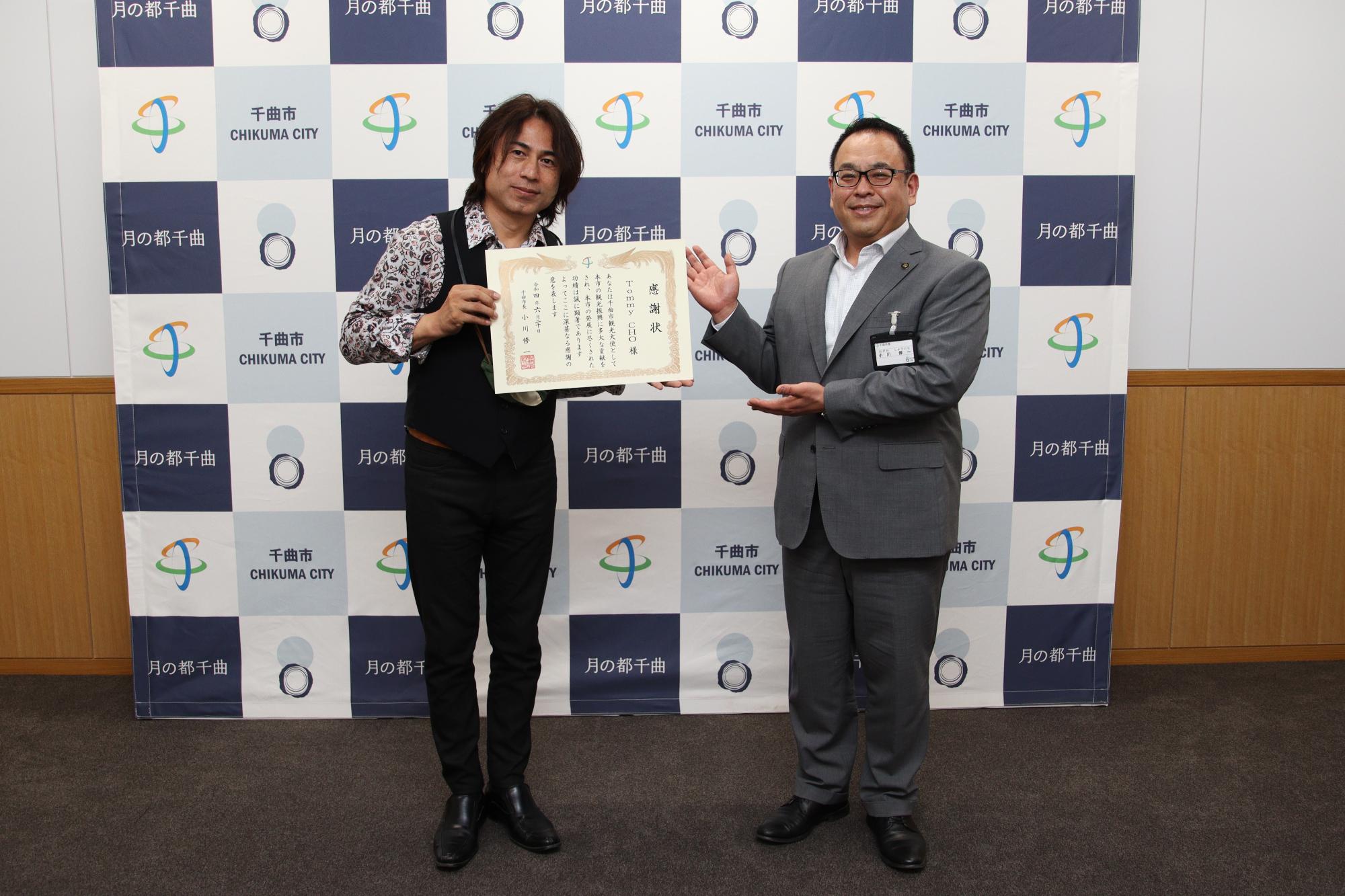 感謝状を手に取るトミーチョウさんと小川修一市長の記念撮影