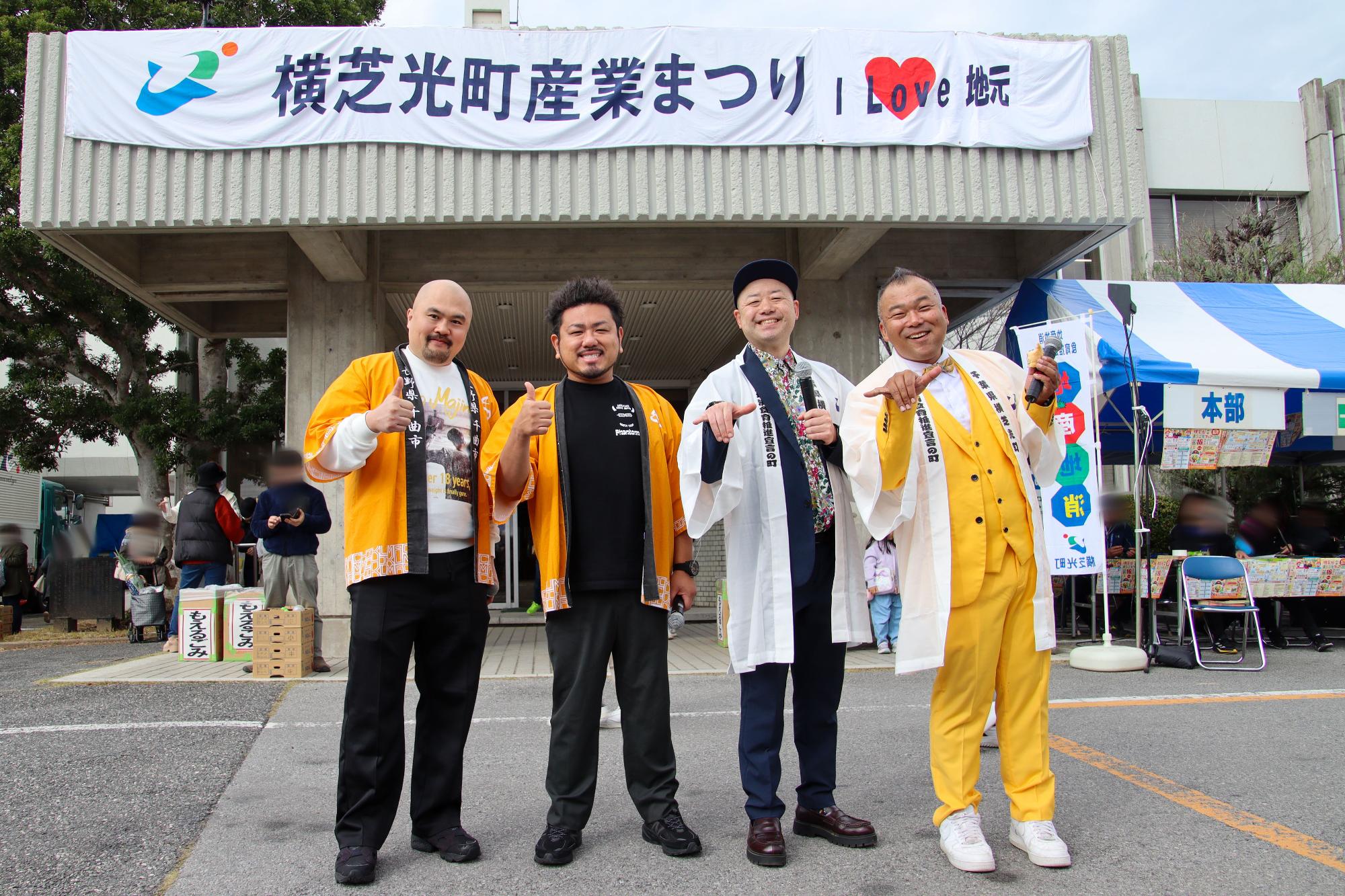 産業まつりの横断幕の下で記念撮影する鬼越トマホーク(左二人)ともぐもぐピーナッツ(右二人)