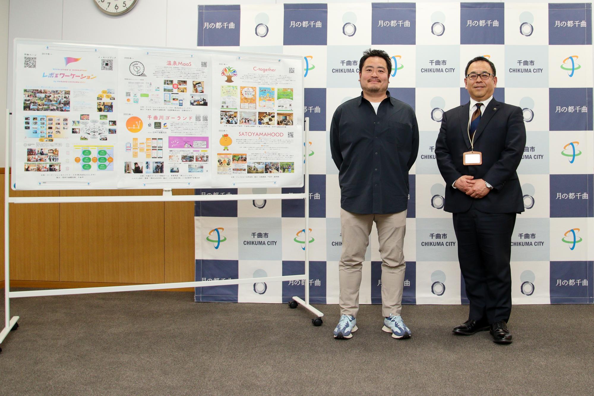田村さんが携わっている事業をまとめた模造紙をバックに記念撮影する田村さん(左)と小川市長(右)