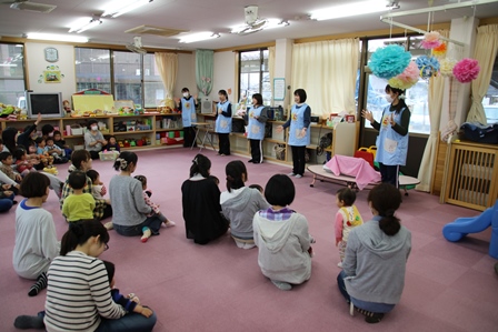 先生たちがレクリエーションをしているのをお母さんの膝の上に子供が座ってみている写真