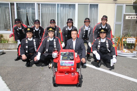 小型ポンプの後ろで市長と団員らが2列に並んで記念撮影をしている写真