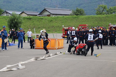周囲に民家などが見える広場で消防団員らが小型ポンプを使い演習を行っている写真