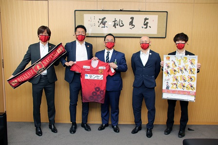 赤いタオルやにユニフォームなどをカメラのほうへ見せながら記念撮影をする市長とボアルース長野の皆さんの写真