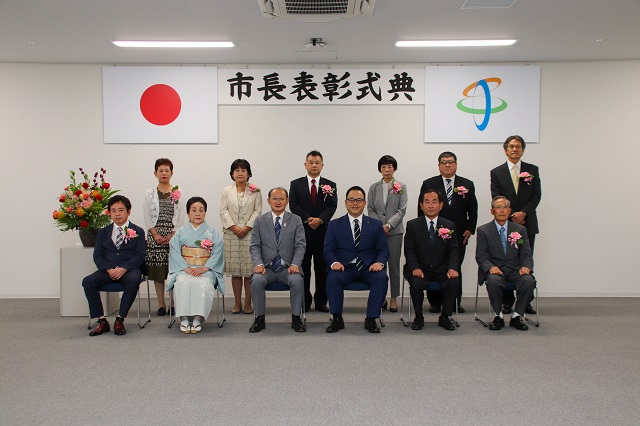 表彰を受けた皆さんが胸元にピンク色の花飾りをつけて記念撮影をしている写真