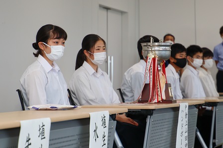 机の上にトロフィーがおかれ、生徒たちが着席している写真