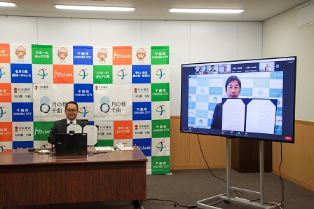 画面に映る長野地域連携中枢都市圏の8市町村長と並び宣言書を披露する小川市長