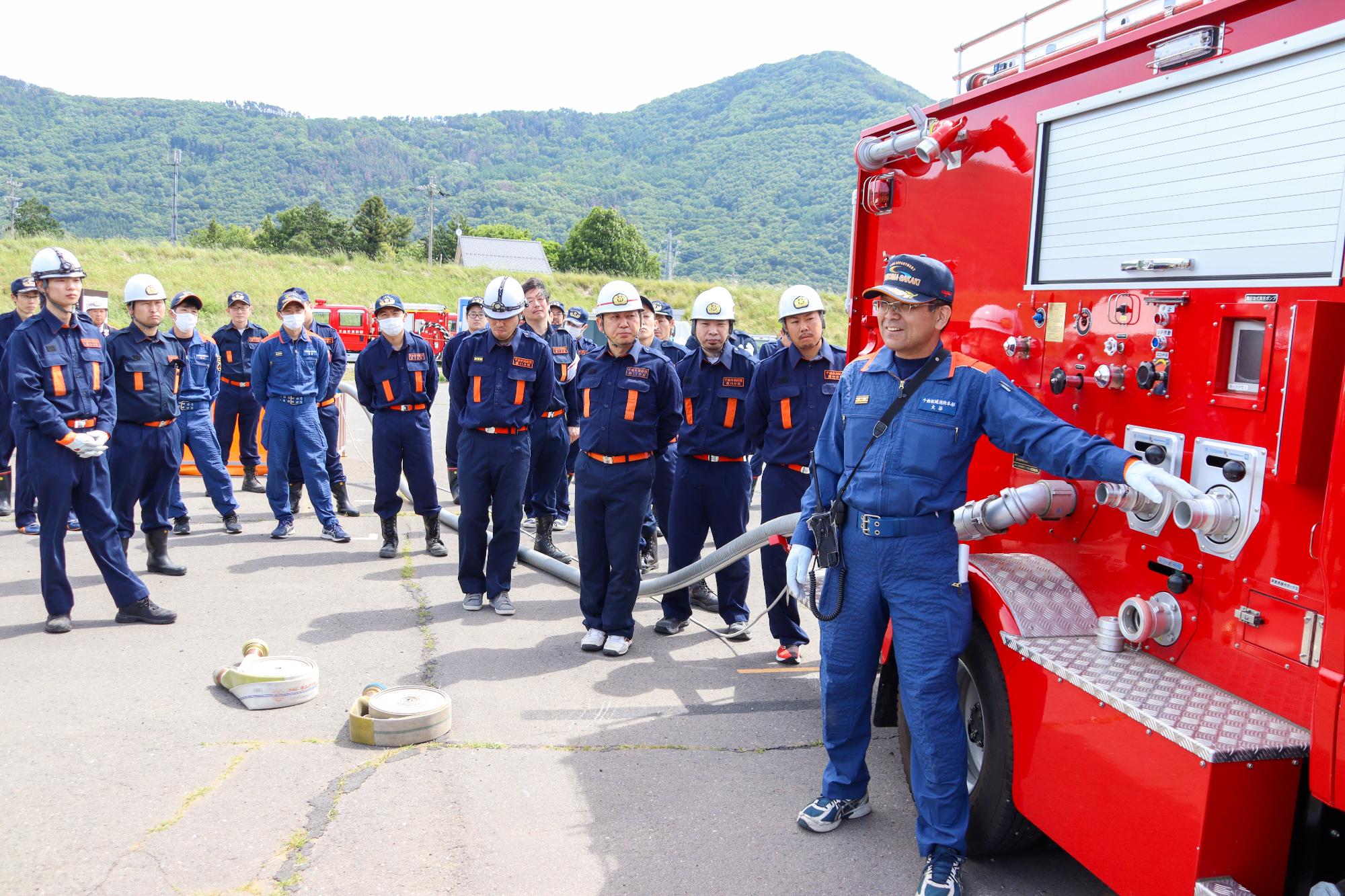 ポンプ車の横で消防本部職員の説明を真剣に聞く消防団員18人