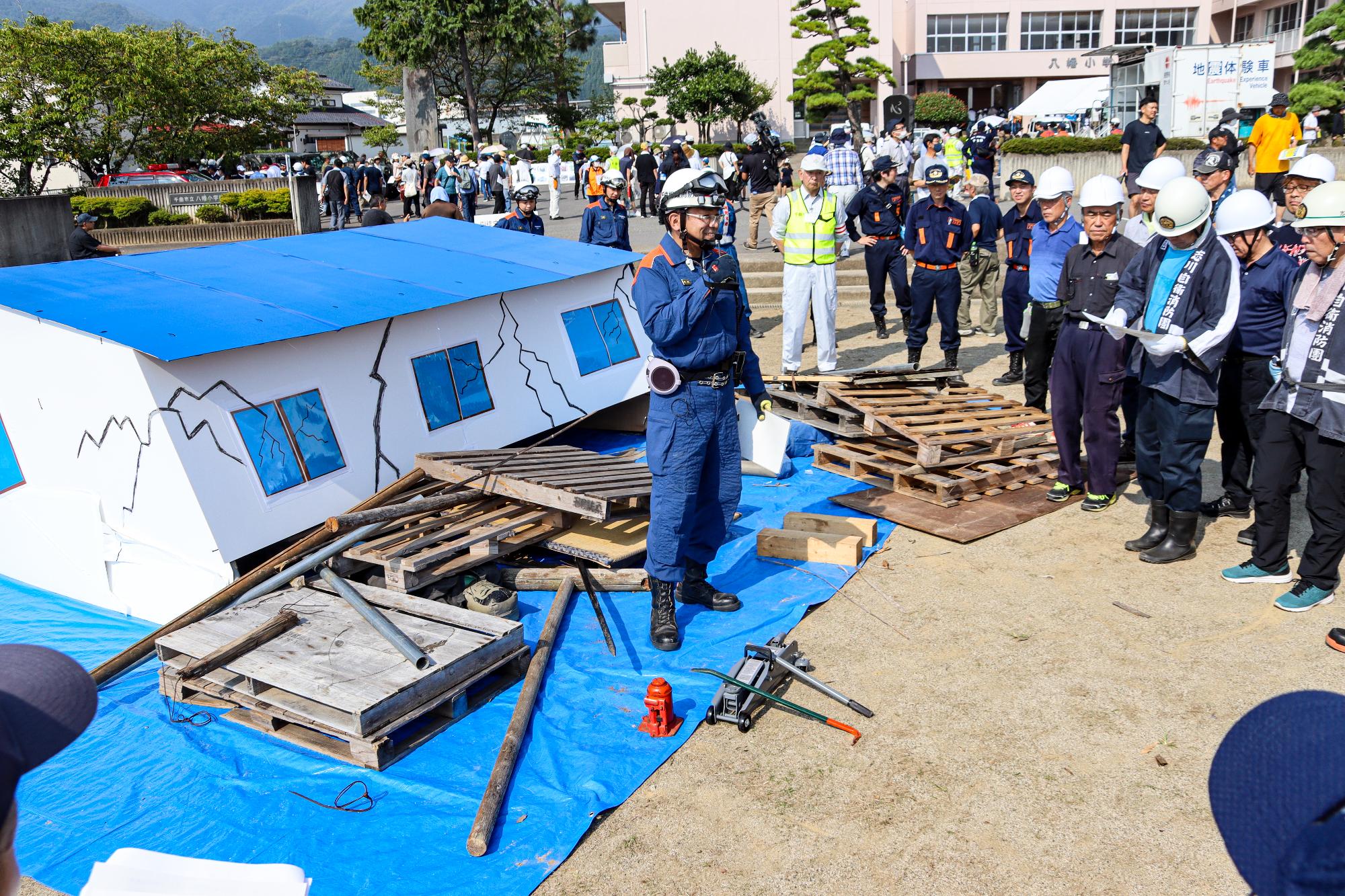 倒壊家屋救出訓練として、倒壊家屋を模した模型の前で説明する消防署員と説明を聞く参加者約15人