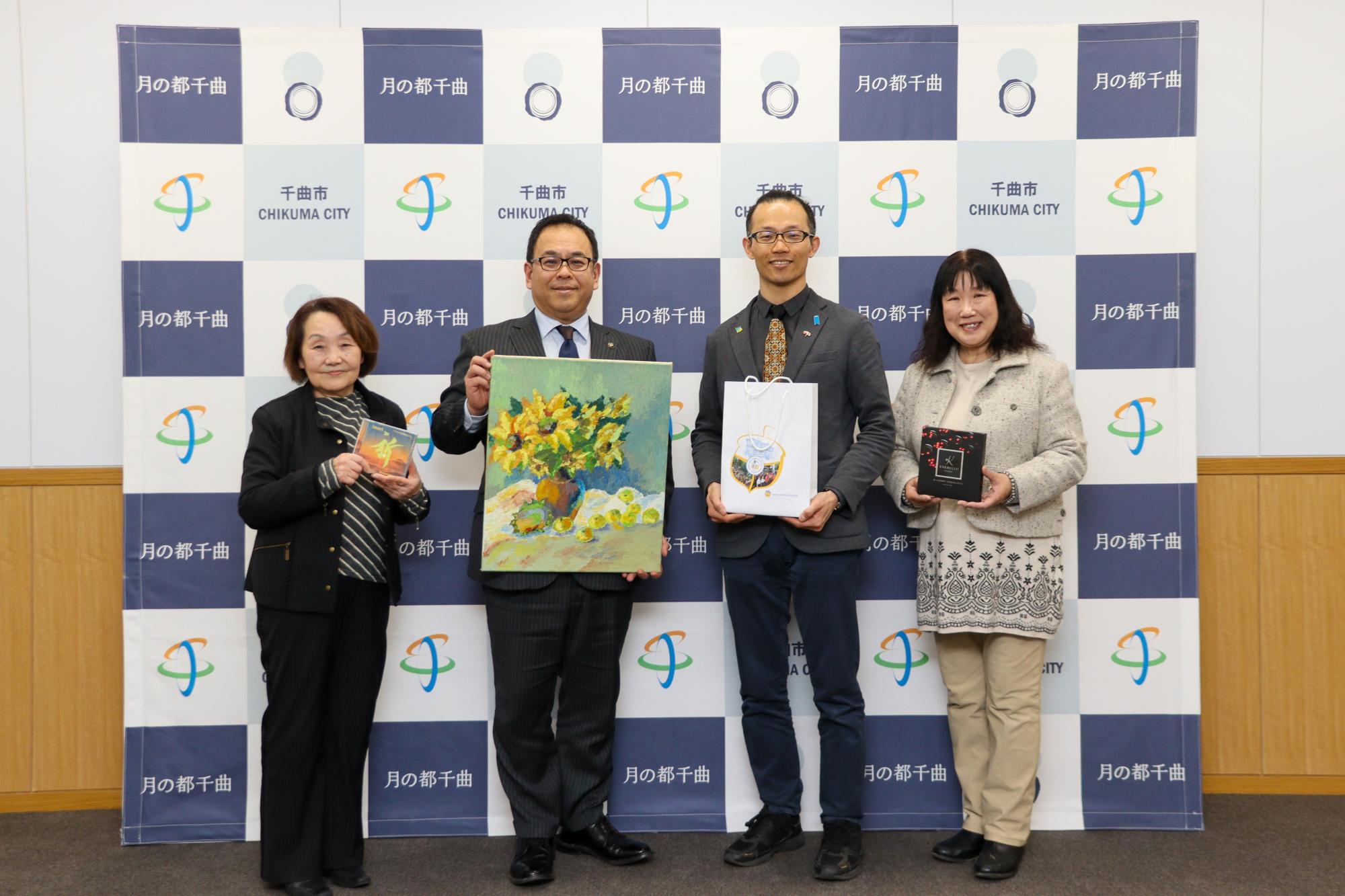 ウクライナの音楽CD、ひまわりの絵、紙袋、チョコレートを持って記念撮影する4人(左から松田教育委員、小川市長、坂本さん、ウクライナの子ども達を支える会の岩田さん)