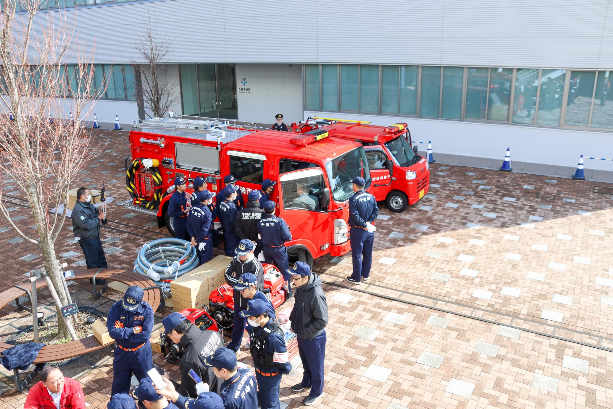 2つの男性の集まりがポンプと車両の周りに集まり、それぞれの使い方の説明を熱心に聞いている様子