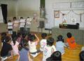 手に画用紙を持って前方に並んでいる子どもたちと、座って見ている子どもたちの写真