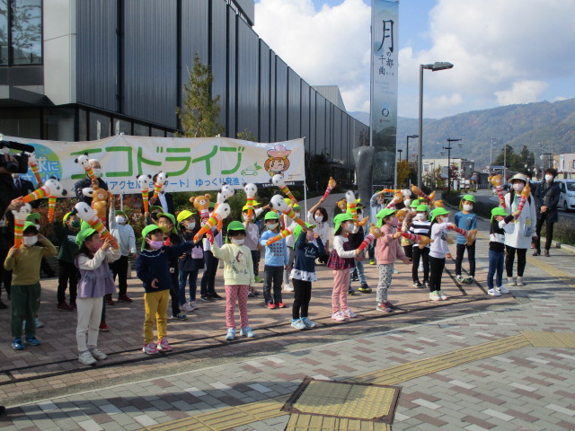 緑色の帽子を被った園児たちが動物のキャラクターがついたスティックを持ち車道に向かって手を振っている写真
