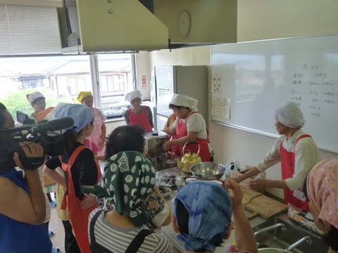 エプロンと三角巾をつけた参加者たちが調理室に集まり料理を教えてもらっている写真