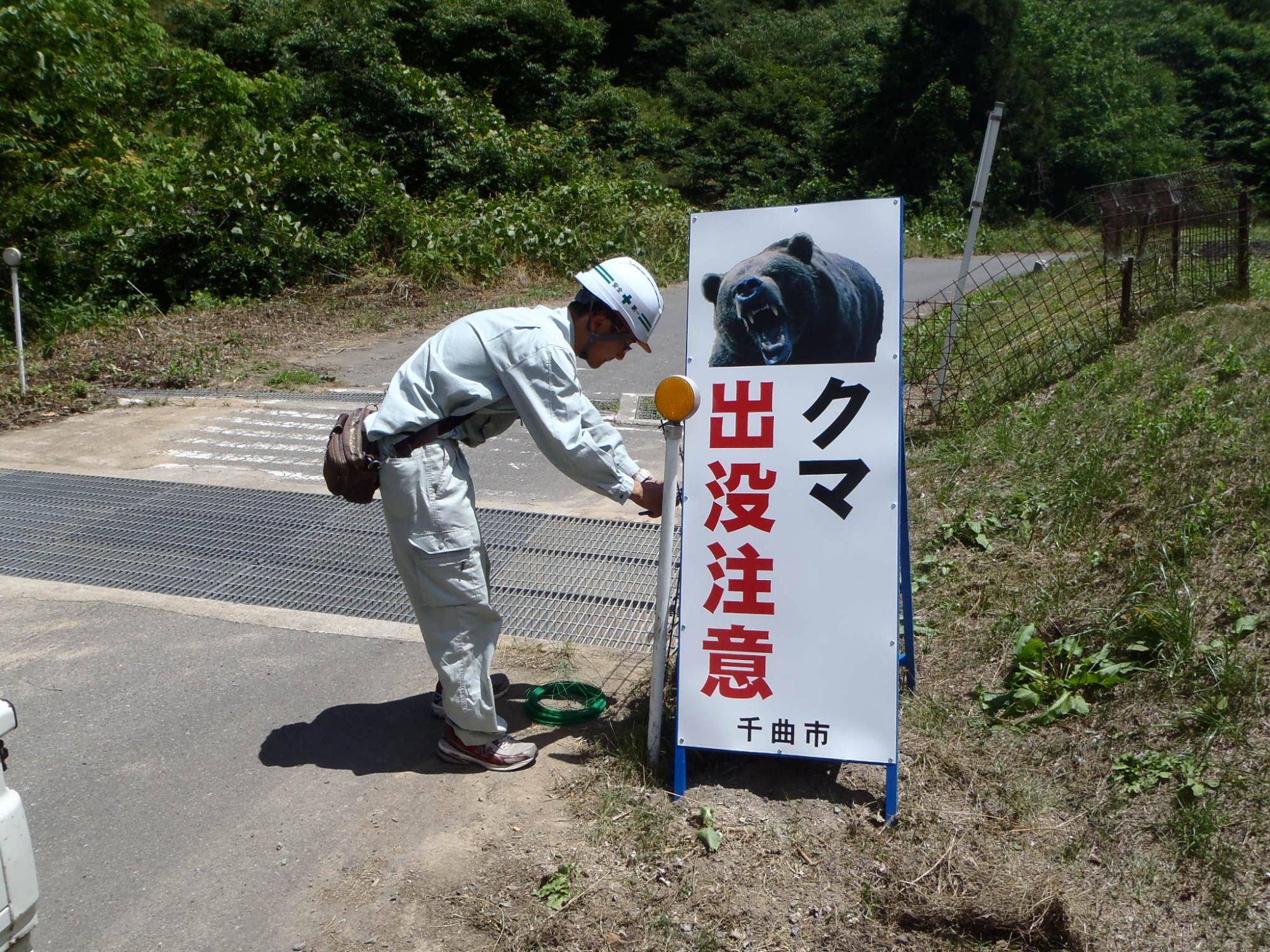 ヘルメットをかぶった作業服姿の男性が、「クマ出没注意」と書かれた看板を設置している写真