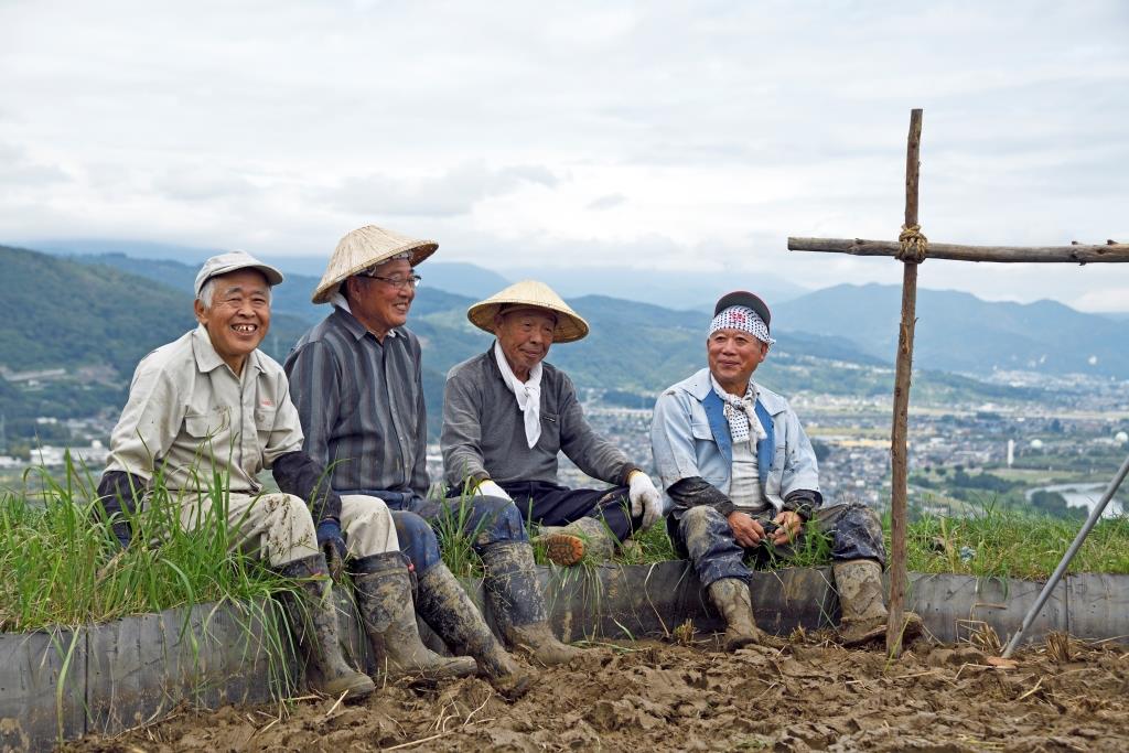 田んぼの端で、4人のお年寄りの男性が笑顔で座っている写真