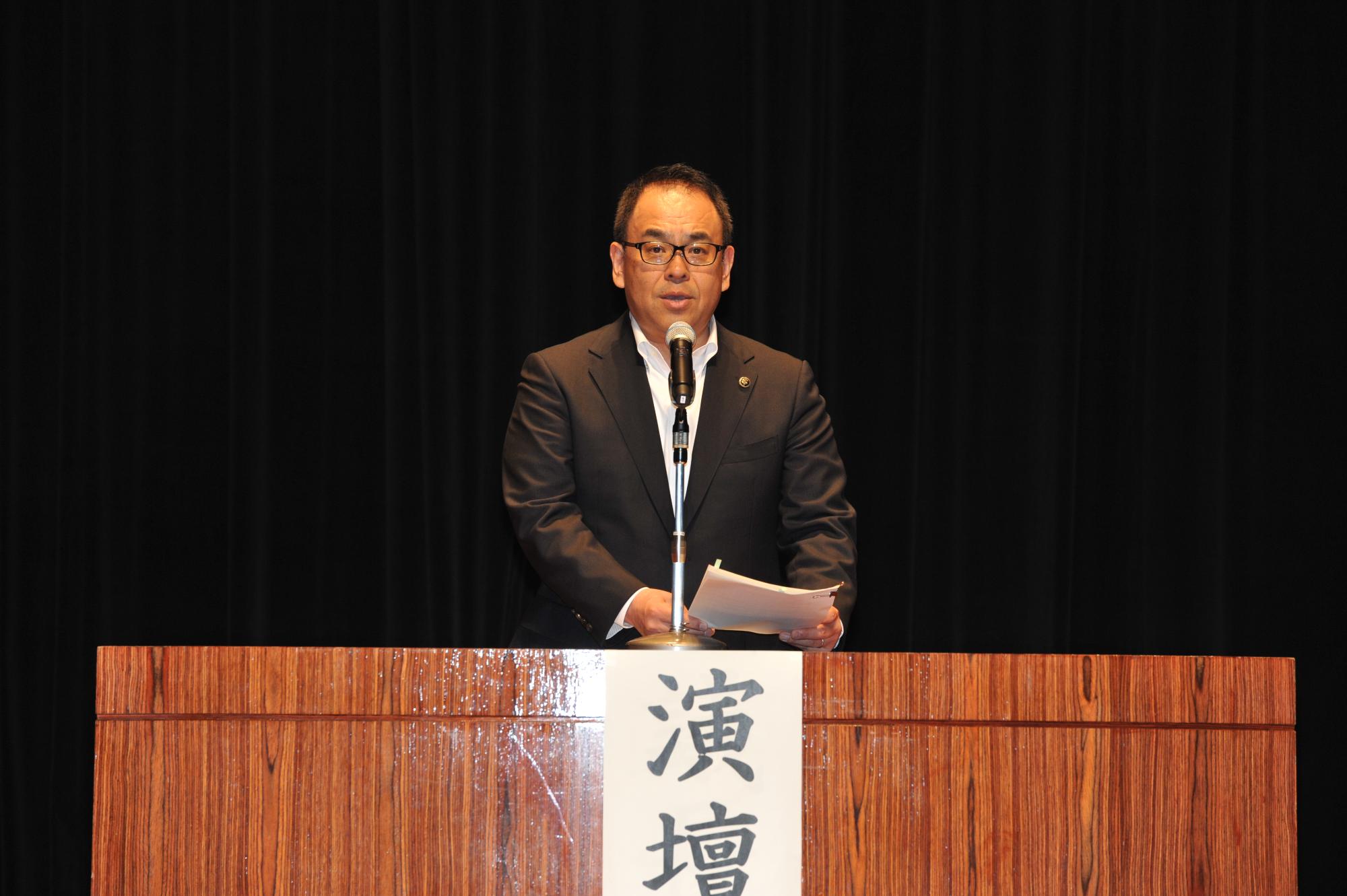 総会で演壇に立った市長がマイクを手に持ち話している写真