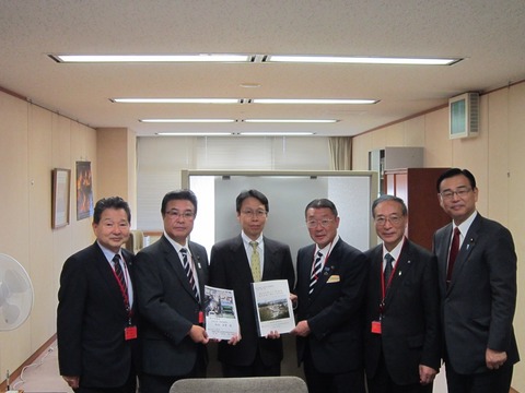冊子を手に持ち、部屋の真ん中に並んで立つ市長と和田総括審議官、および関係者のみなさんの写真